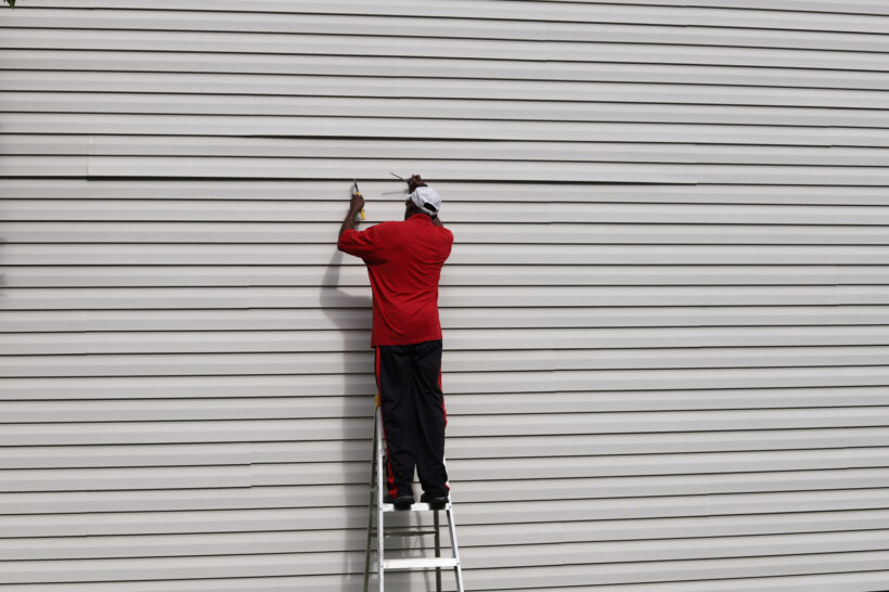 An African-American man on a ladder and fixing vinyl siding on a house, siding contractors East Peoria IL, siding company East Peoria IL, vinyl siding East Peoria IL, siding installation East Peoria IL, siding replacement East Peoria IL, siding contractors near East Peoria IL, house siding contractors East Peoria IL, exterior remodeling East Peoria IL, soffit and fascia East Peoria IL, vinyl siding contractors East Peoria IL, siding repair East Peoria IL, home siding replacement East Peoria IL, insulated siding East Peoria IL, residential siding contractor East Peoria IL, low maintenance siding East Peoria IL, siding installers East Peoria IL, curb appeal siding East Peoria IL, siding estimate East Peoria IL, Norandex siding East Peoria IL, Mastic siding East Peoria IL, siding services East Peoria IL